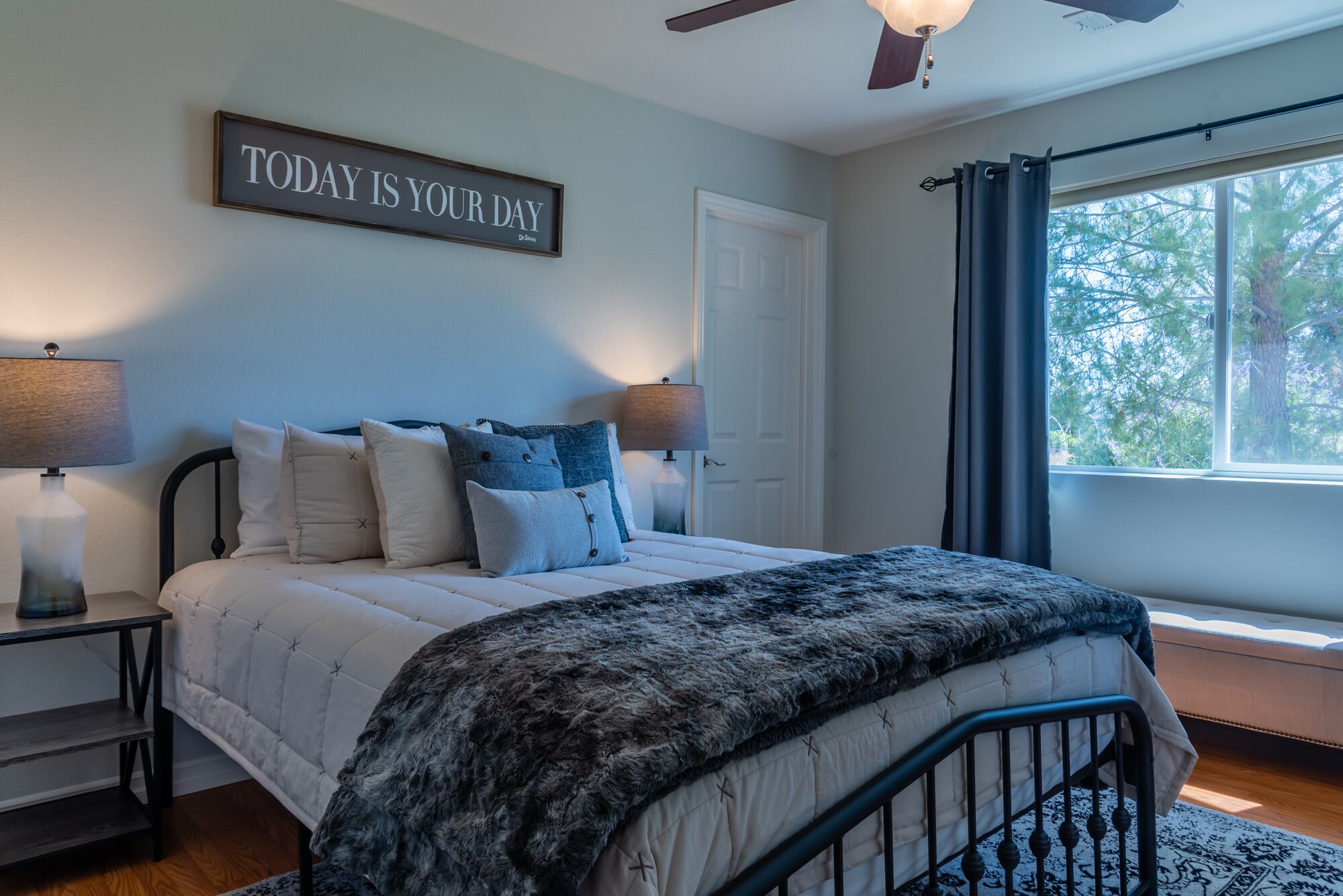 A bedroom with a bed, two bedside tables with lamps, a ceiling fan, a window with curtains, and a motivational sign on the wall.