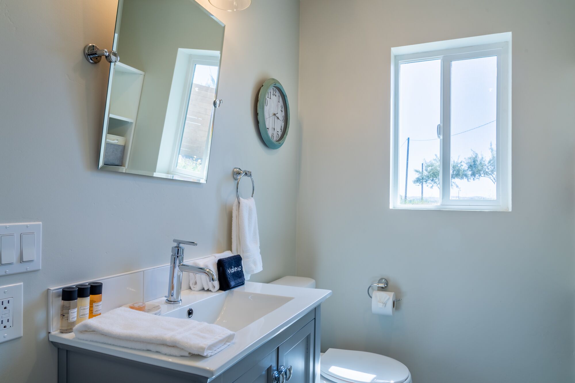 A bathroom with a sink, mirror, clock, and window.