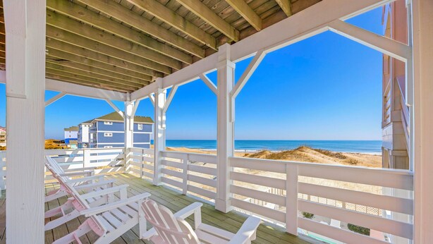 Eine Holzterrasse mit weißen Geländern, die auf einen Strand mit Häusern im Hintergrund blickt.