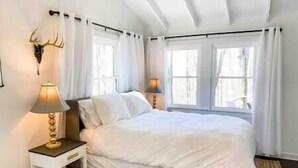 A bedroom with a bed, a nightstand, a lamp, a wall-mounted deer head, and large windows with curtains. - Mountain Retreat: Virginia Foothills (Charlottesville)