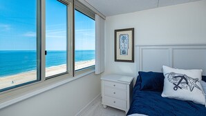 Ein Schlafzimmer mit großem Fenster mit Blick auf einen Strand, ein Bett mit blauem Kopfkissen, ein Nachttisch und ein gerahmtes Kunstwerk an der Wand.