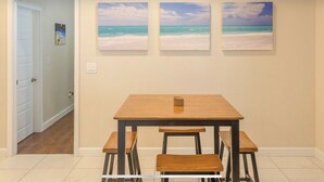 Un coin repas avec une table et des bancs en bois, trois tableaux sur le thème de la plage accrochés au mur, et une porte donnant sur une autre pièce.