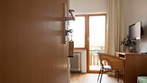 Een hotelkamer met een houten deur, een bord met het nummer 15, een bureau met stoel, een televisie en een raam met gordijnen.
