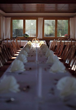 Une longue table à manger, dressée pour un repas, avec des chaises de chaque côté, de grandes fenêtres avec des rideaux et une vue sur la verdure à l’extérieur.