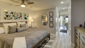 A bedroom with a bed, a ceiling fan, a nightstand, a lamp, a mirror, and a hallway leading to another room. - Spectacular Oceanfront Views! Steps to the Sand and Coligny (Hilton Head Island)