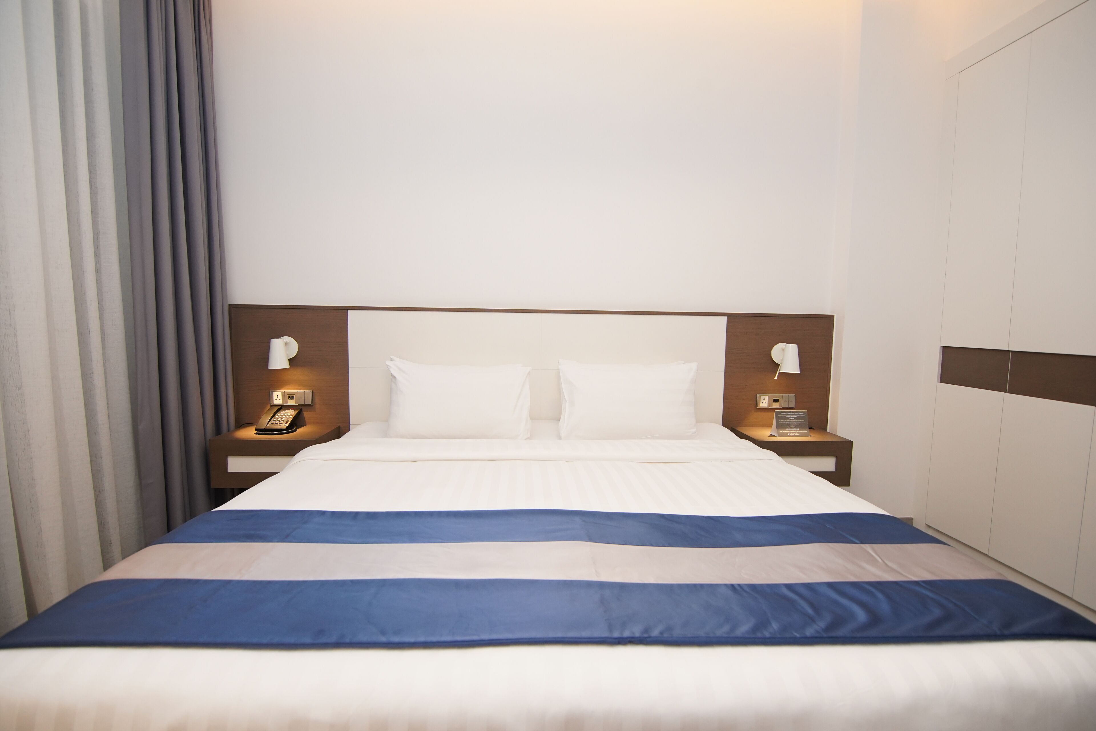 A hotel room with a large bed, two bedside tables with lamps, a wooden headboard, and a curtain on the left.