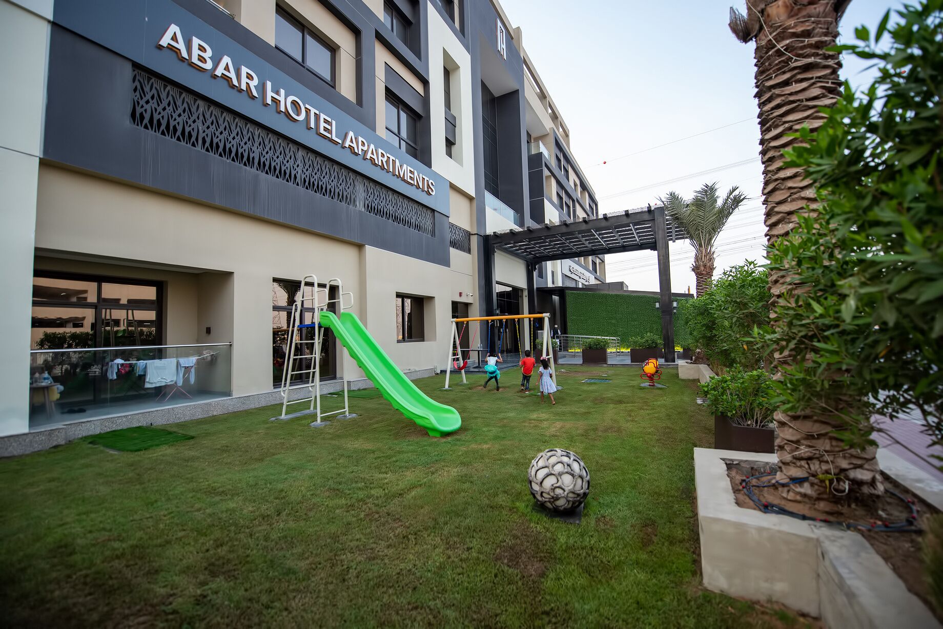 Área externa com parquinho infantil, incluindo escorregador e balanços, em frente ao hotel "ABAR HOTEL APARTMENTS".