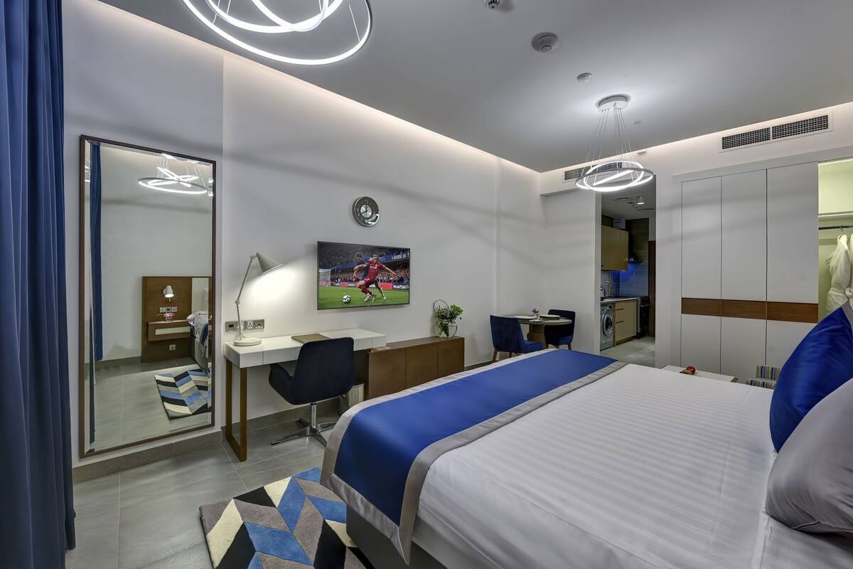 A modern hotel room with a bed, desk, chair, TV, and a small kitchenette.