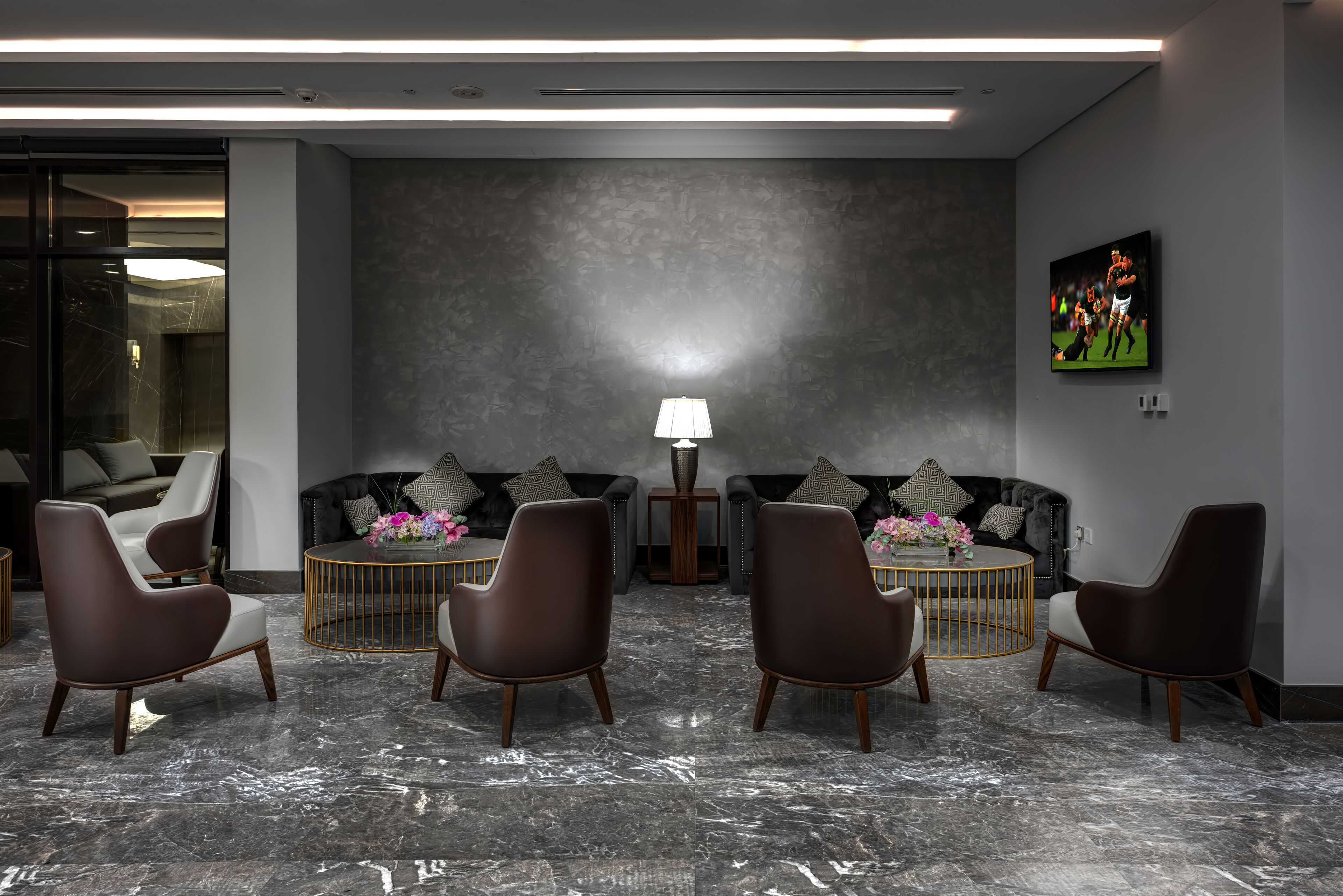 A modern lobby with a marble floor, leather chairs, a round table with a vase of flowers, a lamp, and a TV on the wall.
