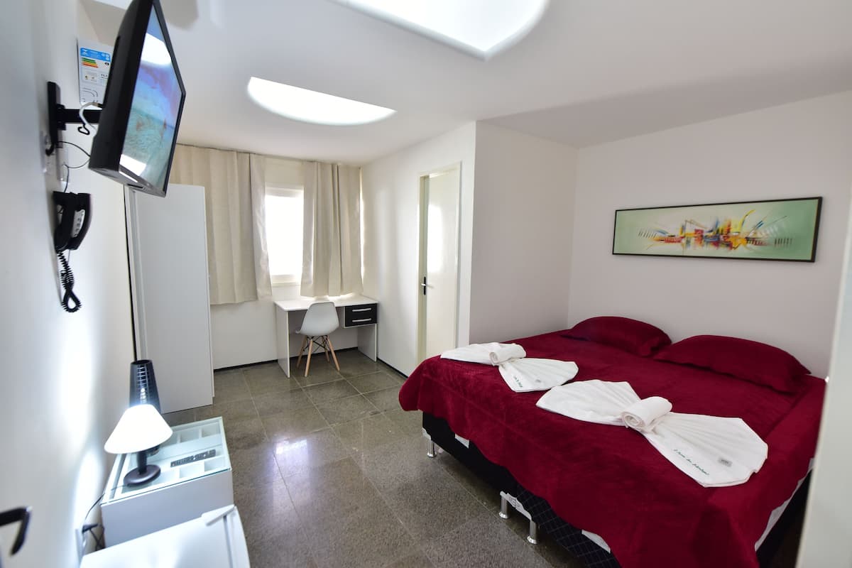 A hotel room with a bed, a desk, a chair, a TV, and a painting on the wall.