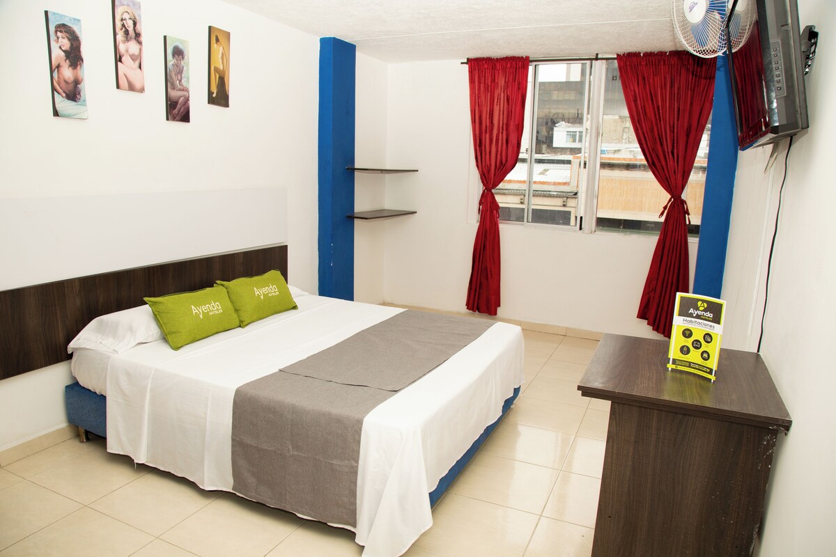 A hotel room with a bed, a desk, a fan, a window with red curtains, and artwork on the wall.