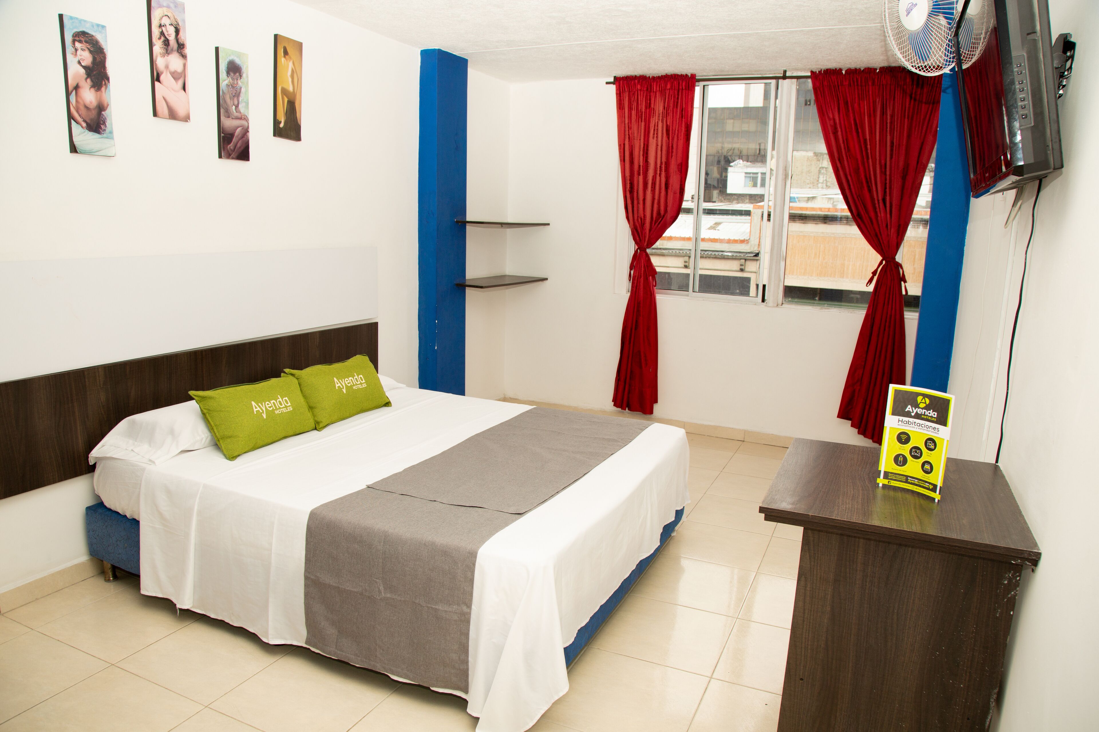A hotel room with a bed, pillows, a desk, a fan, a window with curtains, and a wall with artwork.