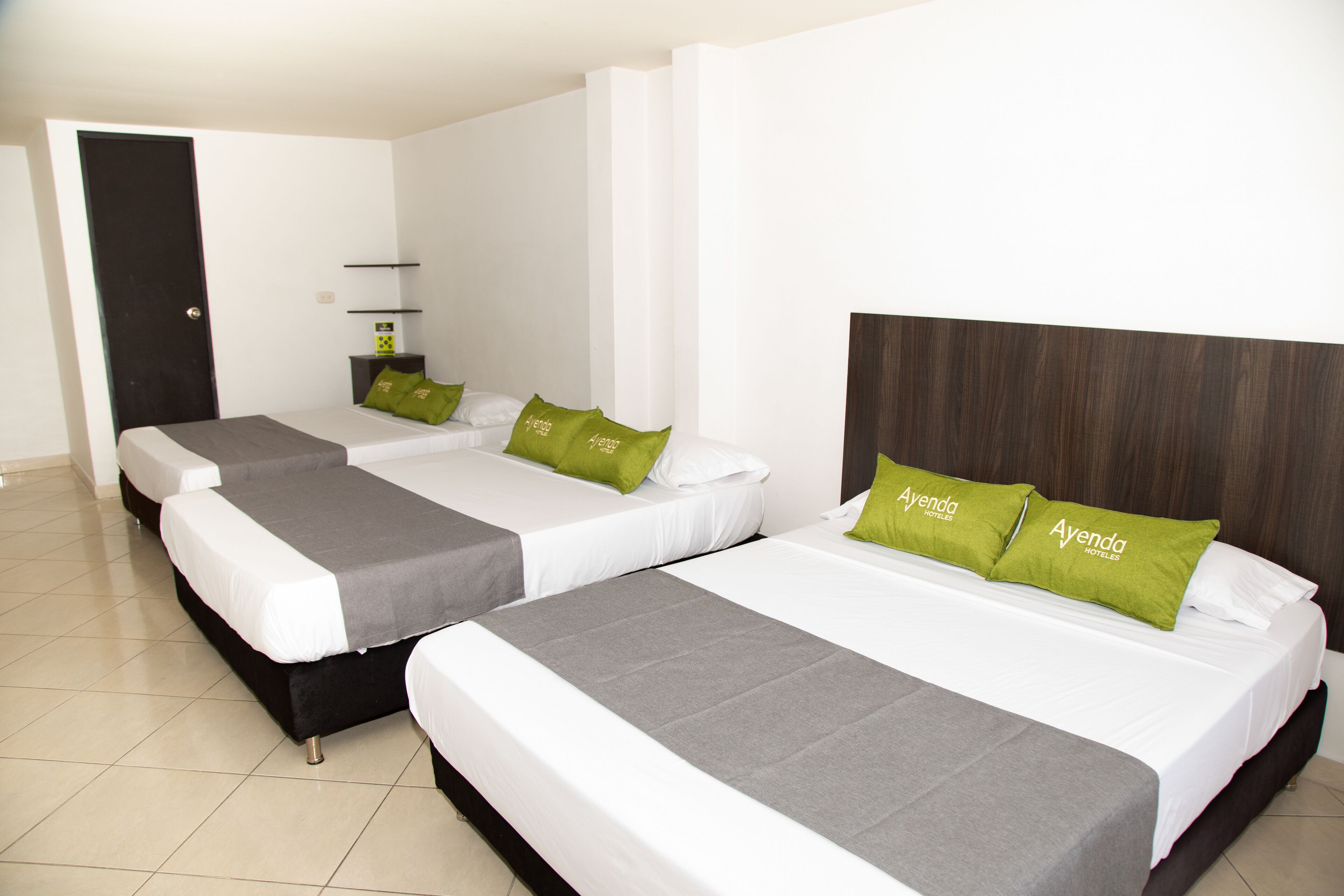 Habitación de hotel con tres camas, cada una con ropa de cama blanca y almohadas verdes, y un cabecero oscuro.