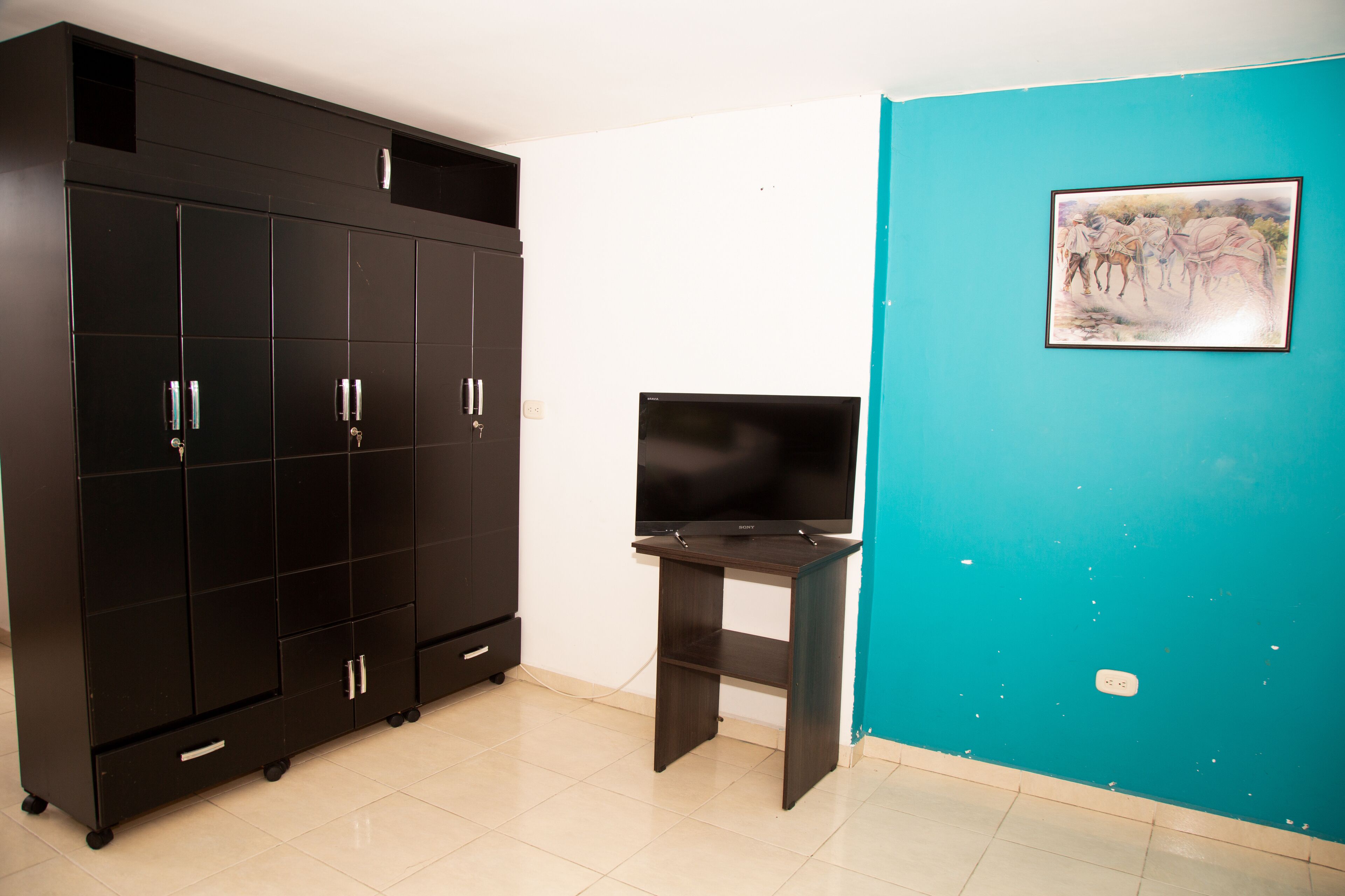 Habitació amb un armari negre gran, televisió de pantalla plana sobre un suport i una paret color turquesa amb un quadre encapçalat.