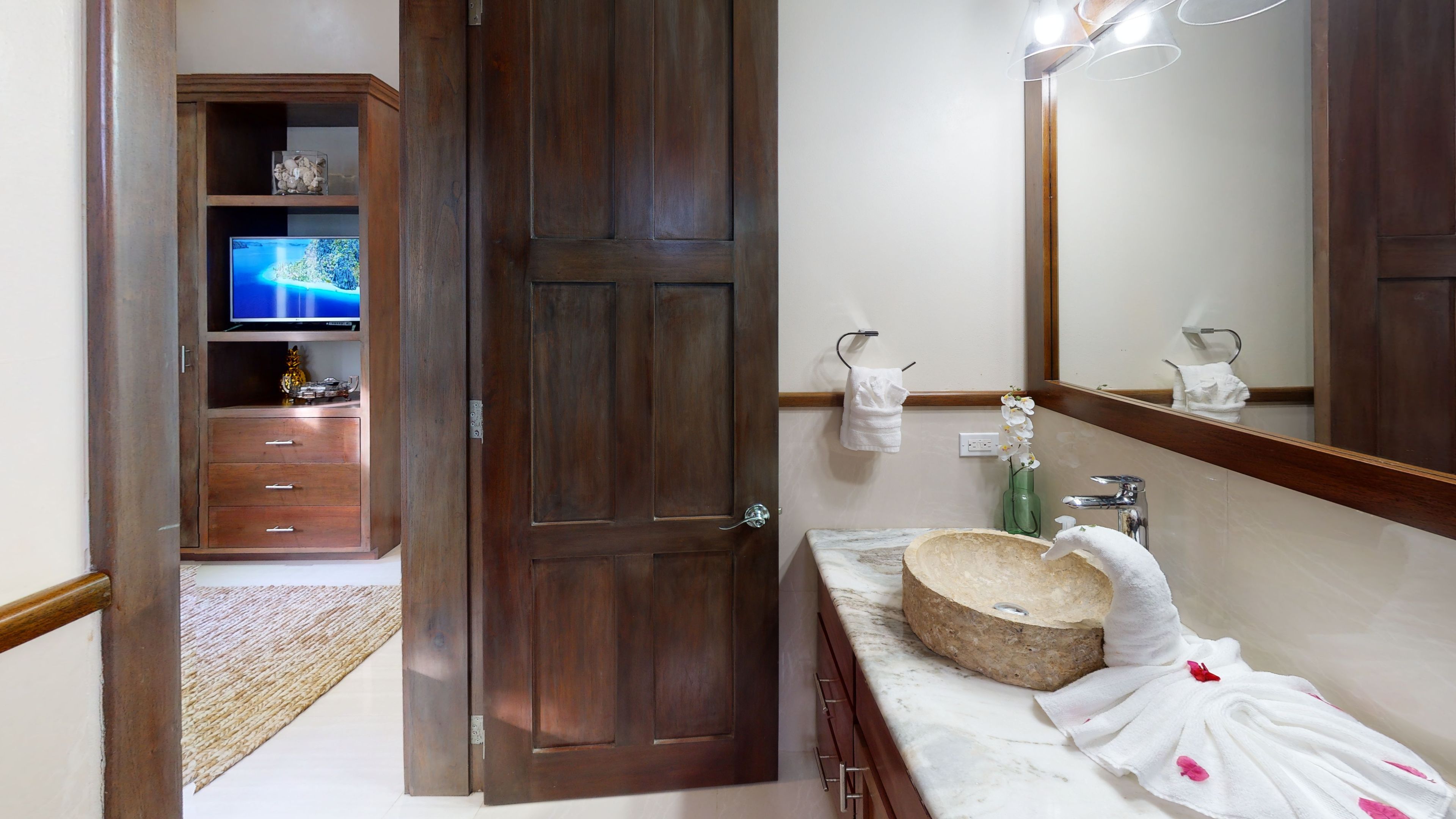 A bathroom with a wooden door, a marble sink, a mirror, and a television in the adjacent room.