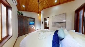 Un dormitorio con techo de madera, un televisor sobre un mueble de madera, una cama con ropa de cama blanca y una almohada azul, y un cuadro en la pared.