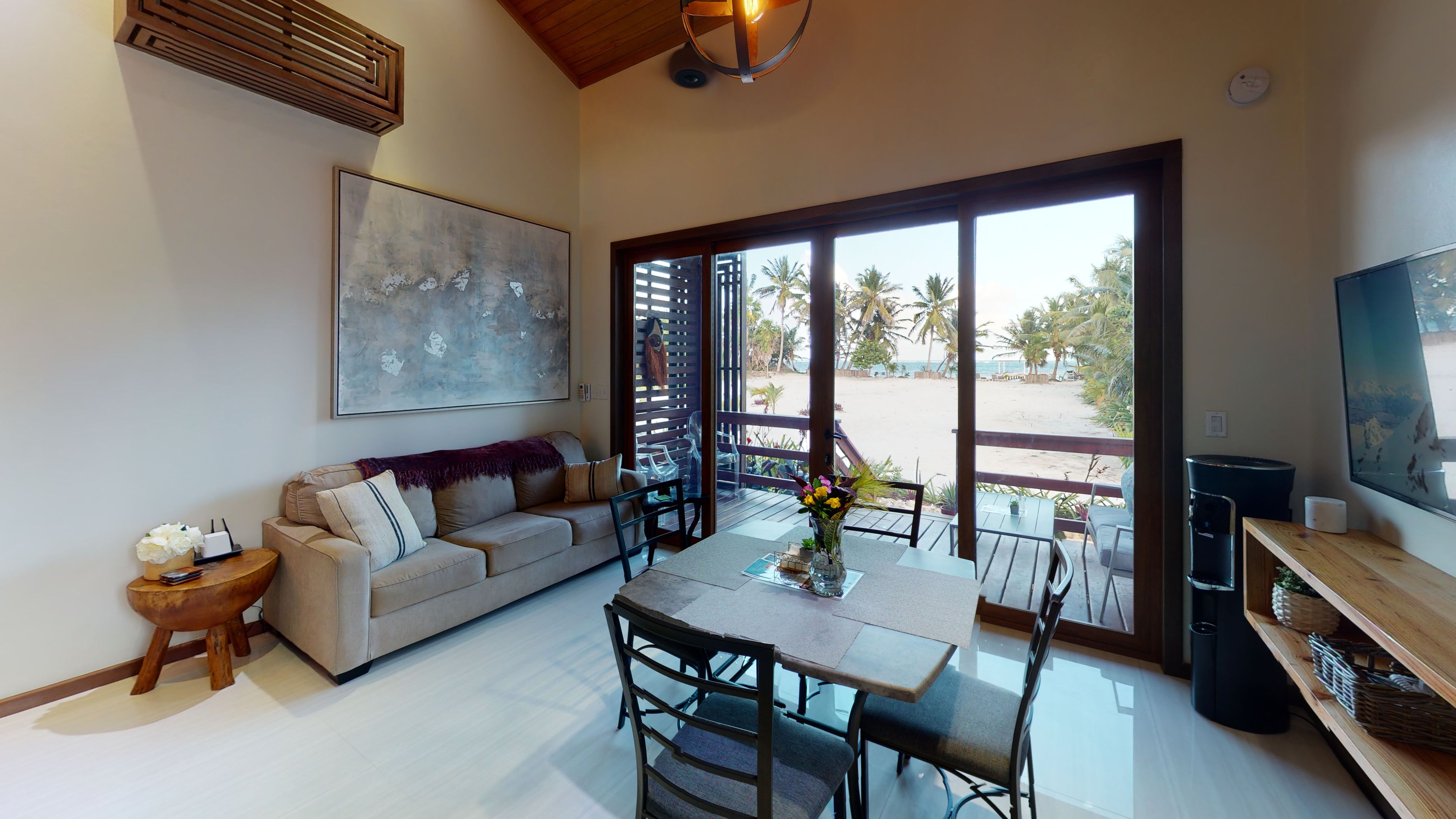 A spacious living area with a sofa, dining table, and chairs, a large abstract painting, and a view of a beach through large windows.
