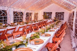 A tented dining area with tables set for a meal, featuring white tablecloths and wooden chairs.