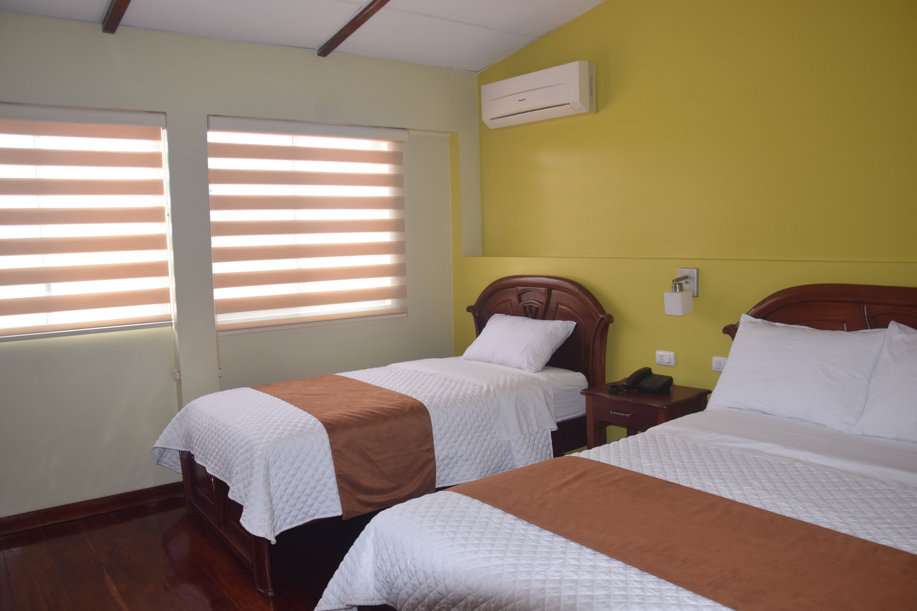 A hotel room with two beds, a wooden headboard, a nightstand, and a wall-mounted air conditioner.