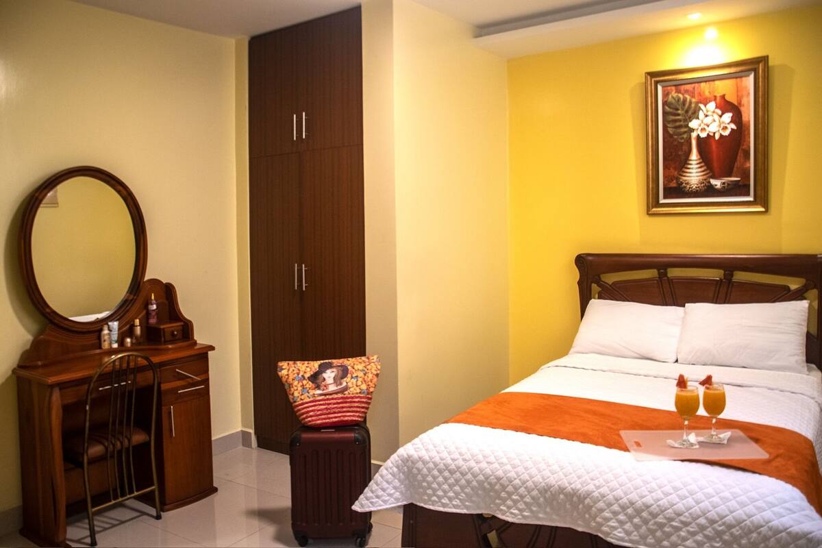 A hotel room with a wooden desk, a large mirror, a wardrobe, a bed with white bedding, and a painting on the wall.