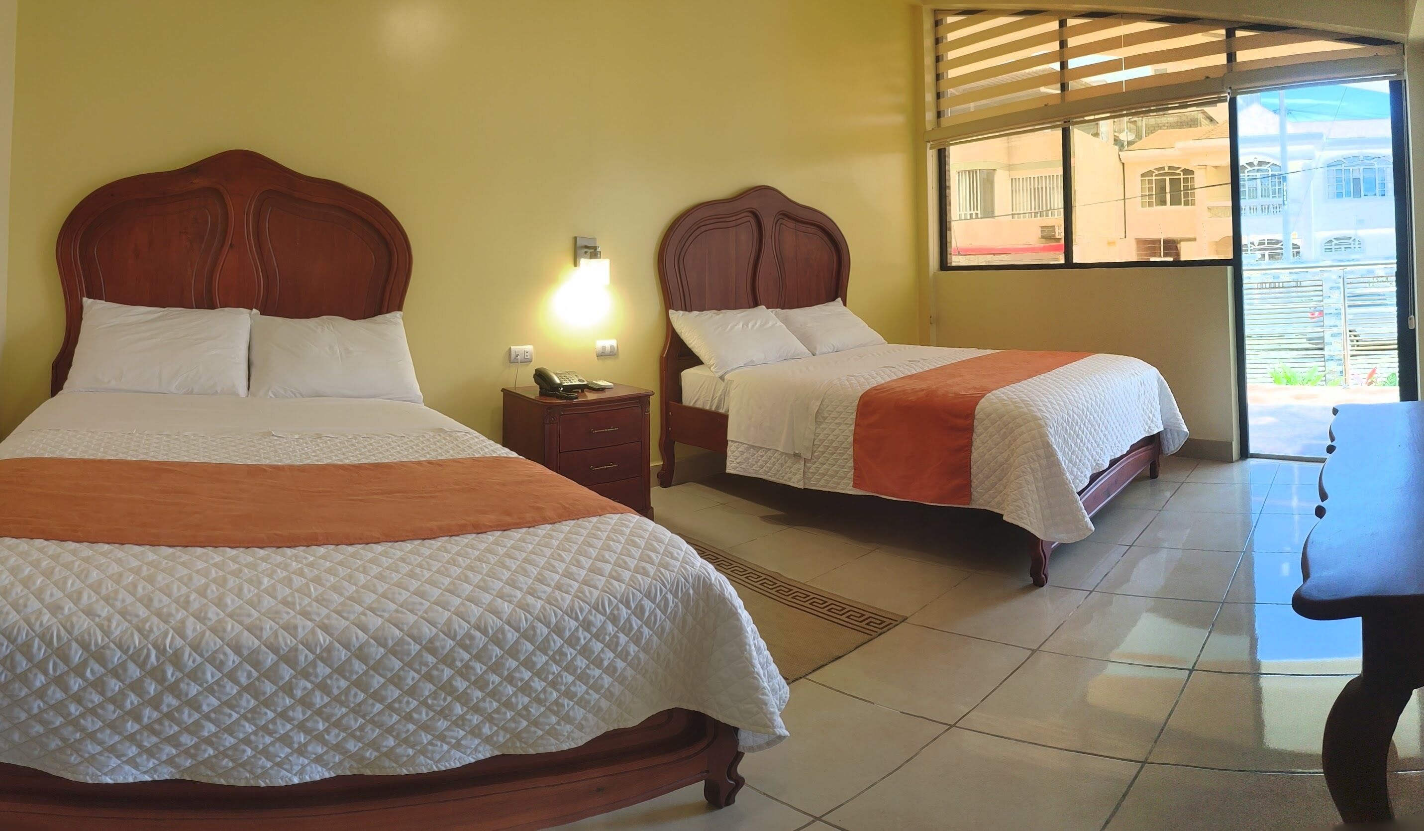 Two double beds with wooden headboards, a bedside table with a lamp, and a view of the outside through a window.
