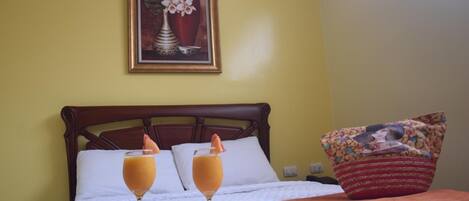 Habitación de hotel con una cama, dos bebidas en una bandeja, un cuadro en la pared y un cojín con estampado floral.