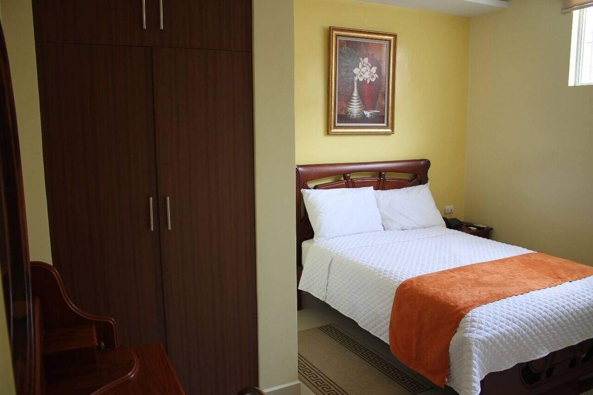 A hotel room with a bed, a wooden wardrobe, and a framed artwork on the wall.
