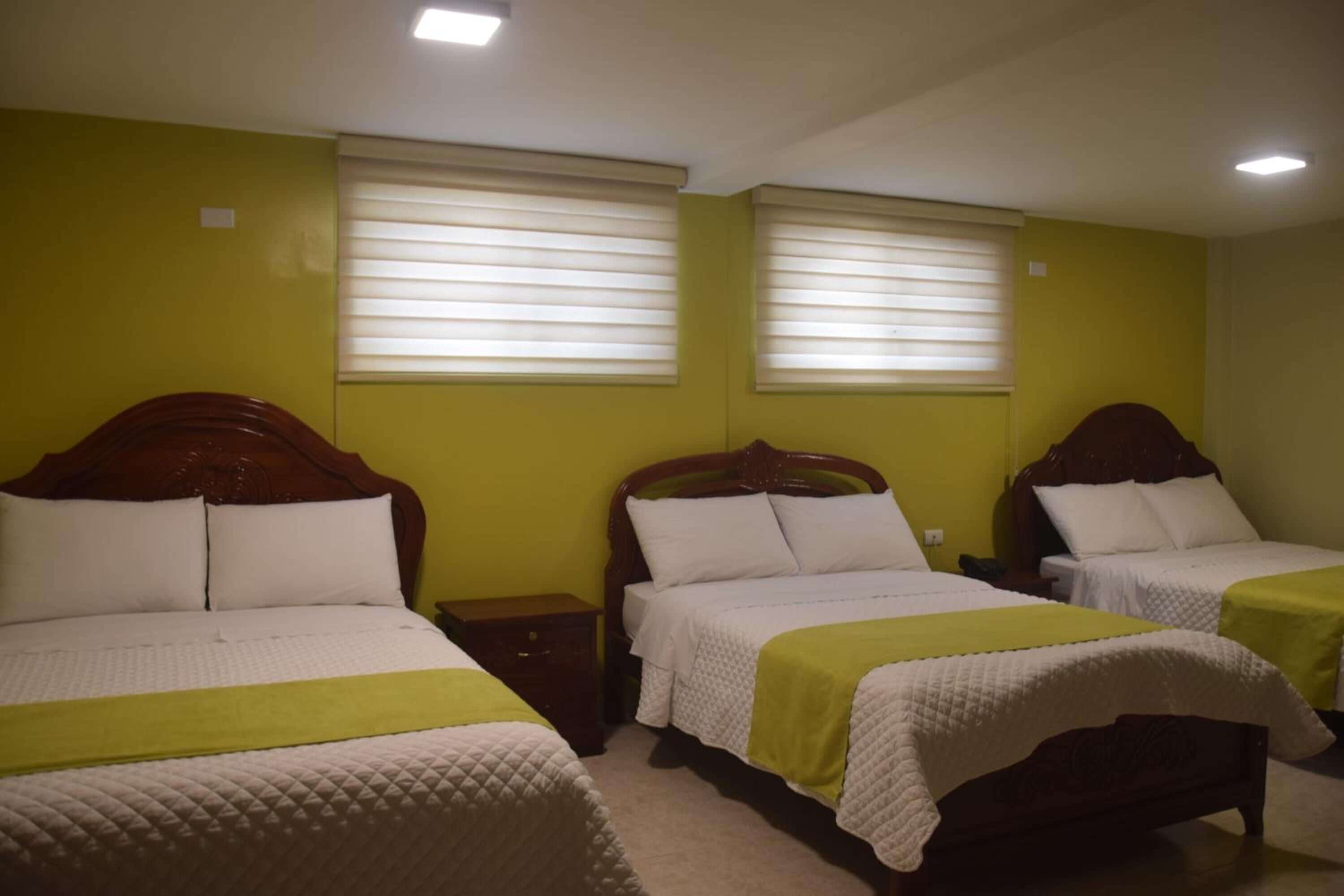 A hotel room with three beds, each with a wooden headboard and white bedding. There are two windows with blinds, and the walls are painted yellow.