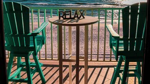 Terraza con dos sillas verdes y una mesa pequeña con un cartel que dice 'RELAX', con vista a una playa al atardecer.