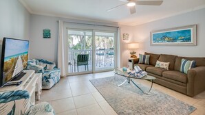 A living room with a sofa, a coffee table, a TV, and a balcony with chairs. - Island Retreat by SeaBreeze Vacation (Holmes Beach)