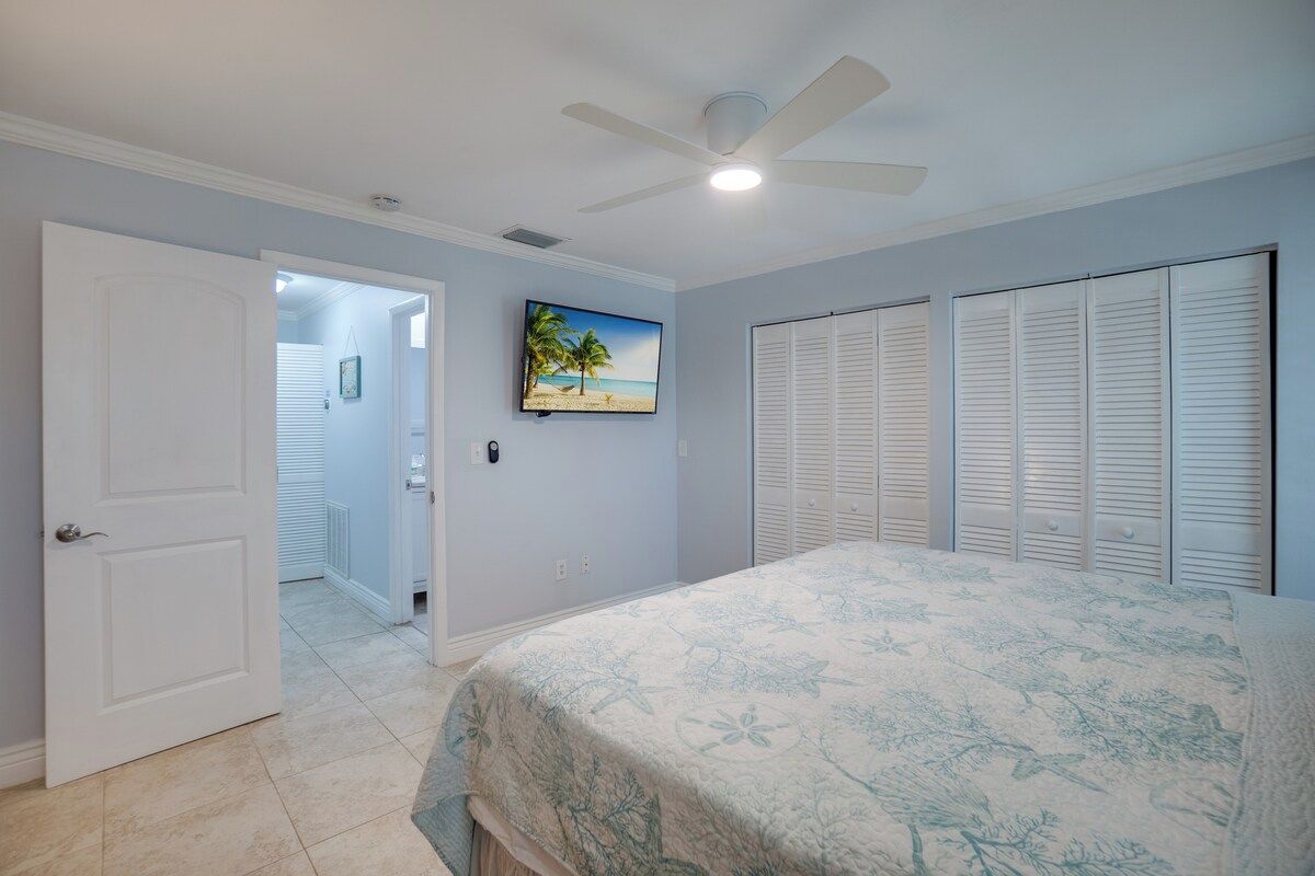 A bedroom with a bed, a ceiling fan, a television, and a door leading to another room.