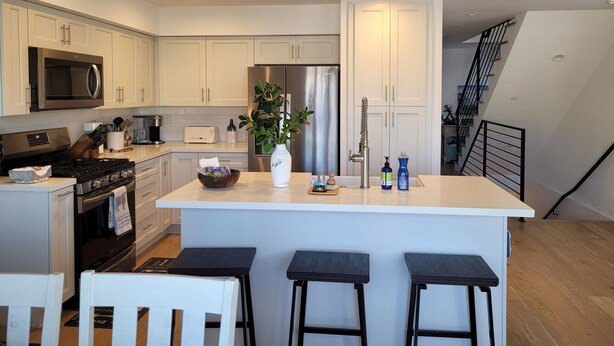A modern kitchen with white cabinets, stainless steel appliances, a central island with stools, and a staircase leading to an upper level. - The ultimate beach getaway charming 3-bdrm 2-bath home steps from a sandy Beach (Oxnard)