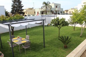 A covered outdoor seating area with a table and chairs, surrounded by a well-maintained lawn and small trees.