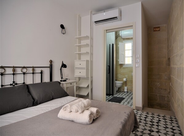 Quarto com cama, mesa de cabeceira com abajur, guarda-roupa branco e banheiro visível por uma porta aberta.
