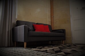 A grey sofa with a red pillow in a room with a tiled floor and a door in the background.
