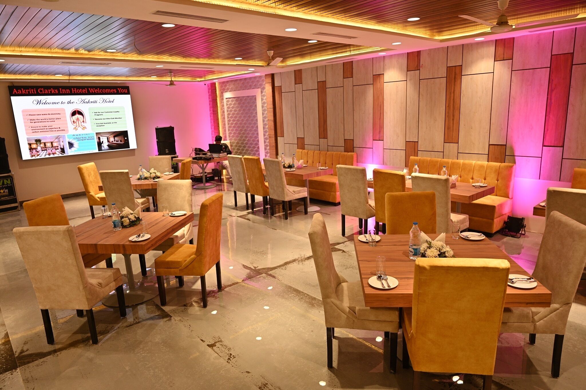Ein Hotelrestaurant mit mehreren Tischen und Stühlen, einem projizierten Bildschirm mit Hotelinformationen und einem modernen Interieur mit Holzwandverkleidungen und Deckenleuchten.