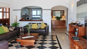 Sala de estar con un sofá de cuero marrón, una mesa de centro de madera y piso a cuadros. Hay una zona de cocina con un microondas y una planta.
