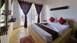 A bedroom with a large bed, a wooden headboard, a red and white pillow set, a bedside table with a bottle, and a view through the window. - Ayla Beach House (Zanzibar Kiwengwa)