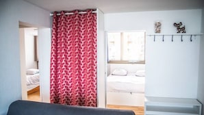 Um quarto com sofá cinza, cortina vermelha estampada, cama branca e prateleira com ganchos fixada na parede.