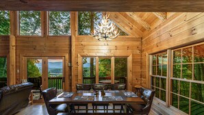 Una baita in legno con zona pranzo, caratterizzata da un grande tavolo e sedie, un lampadario e ampie finestre con vista panoramica.