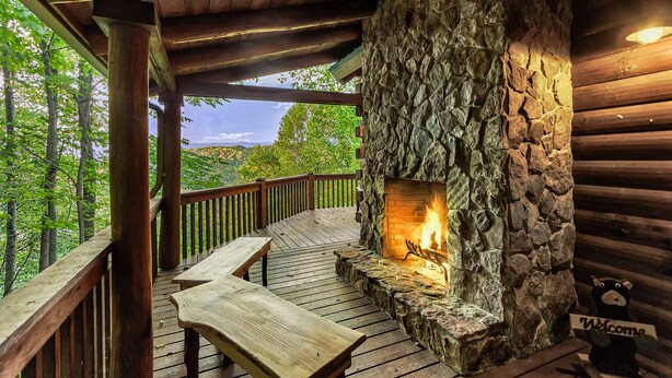 Camino in pietra con fuoco acceso, veranda in legno e vista sulla foresta.