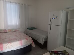 Una habitación pequeña y sencilla con cama, mesita de noche, refrigerador y ventana con cortinas.