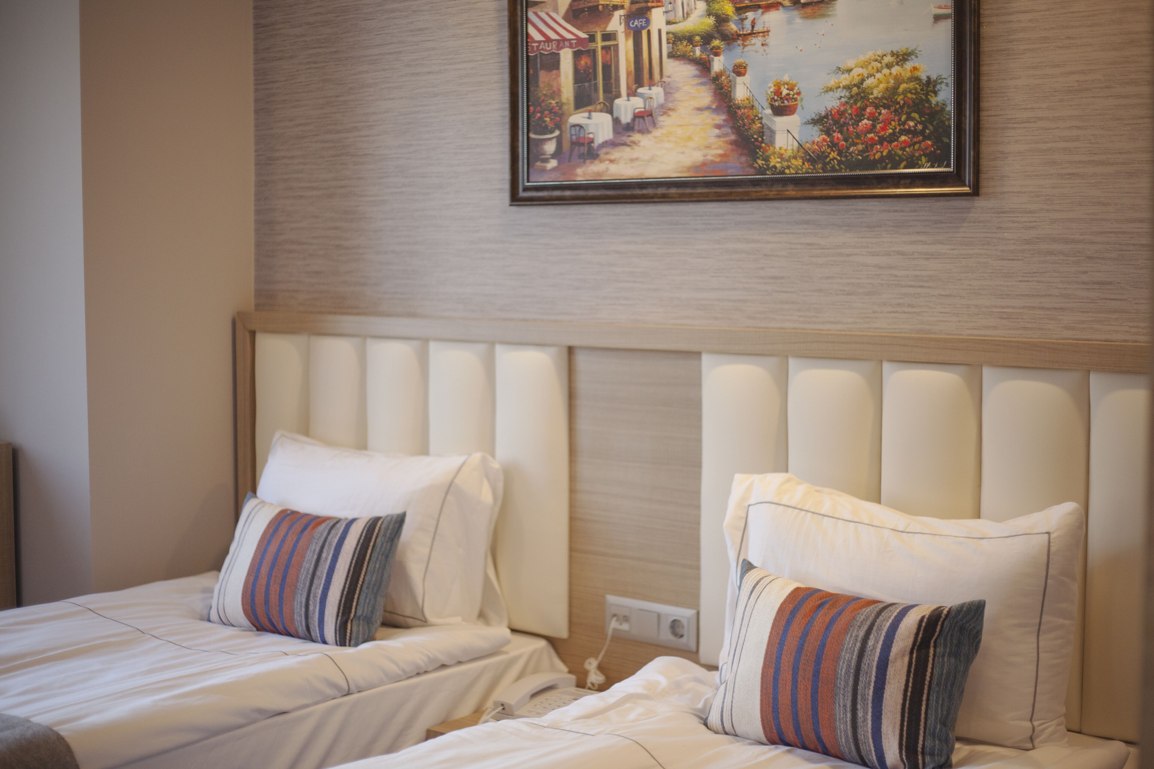 A hotel room with two beds, each with striped pillows, a headboard, and a painting of a street scene above.