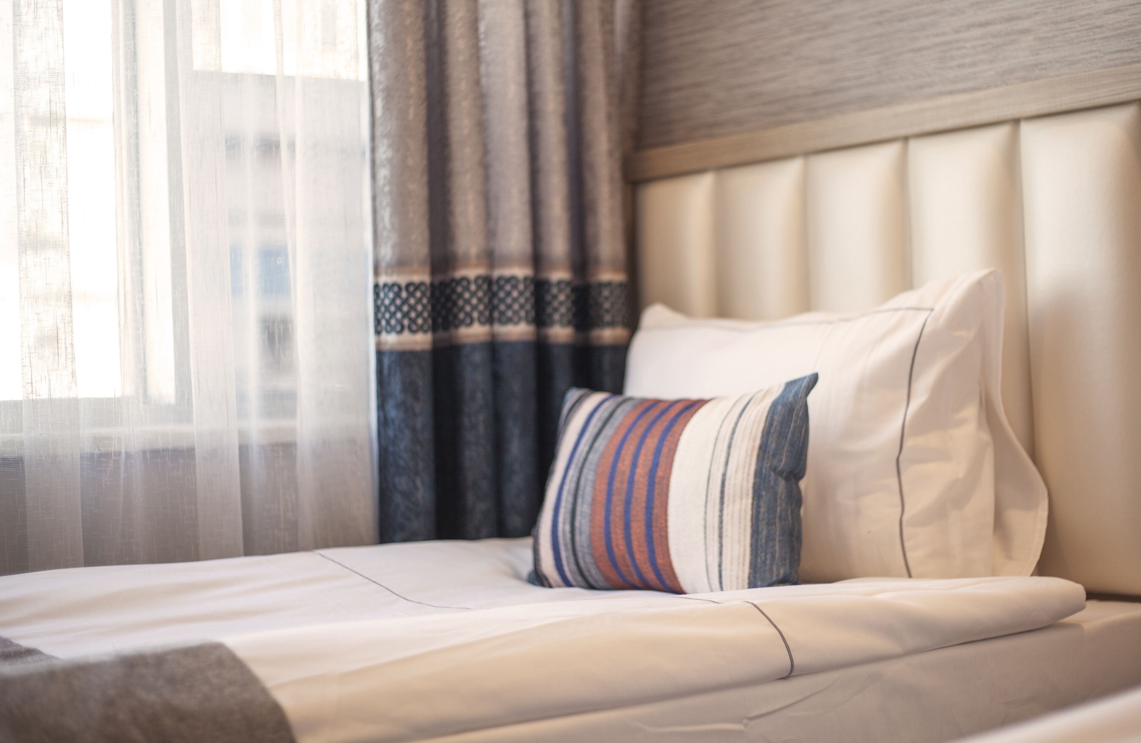 A hotel room with a neatly made bed, a striped pillow, and a window with sheer curtains.