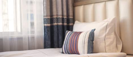 A hotel room with a neatly made bed, a striped pillow, and a window with sheer curtains.