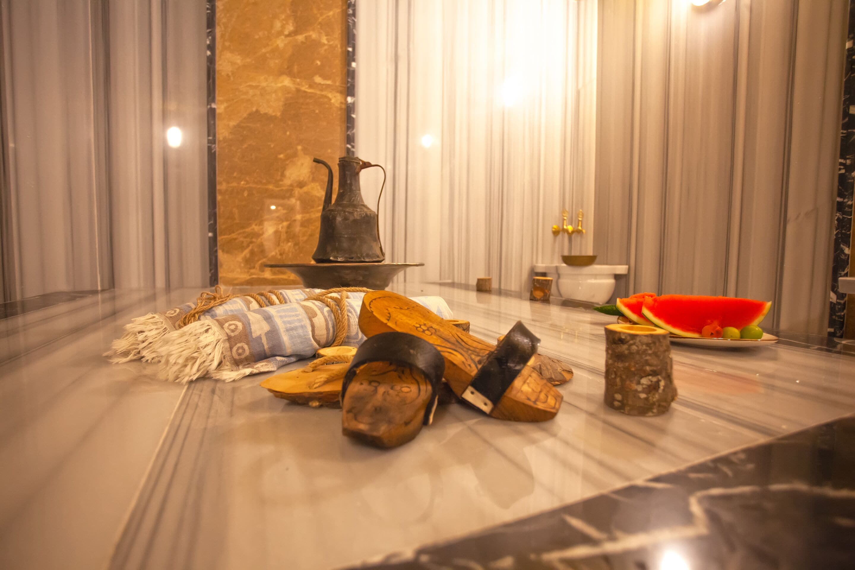 A room with a marble floor, a wooden bench, a small table with a bowl of fruit, and a decorative vase.