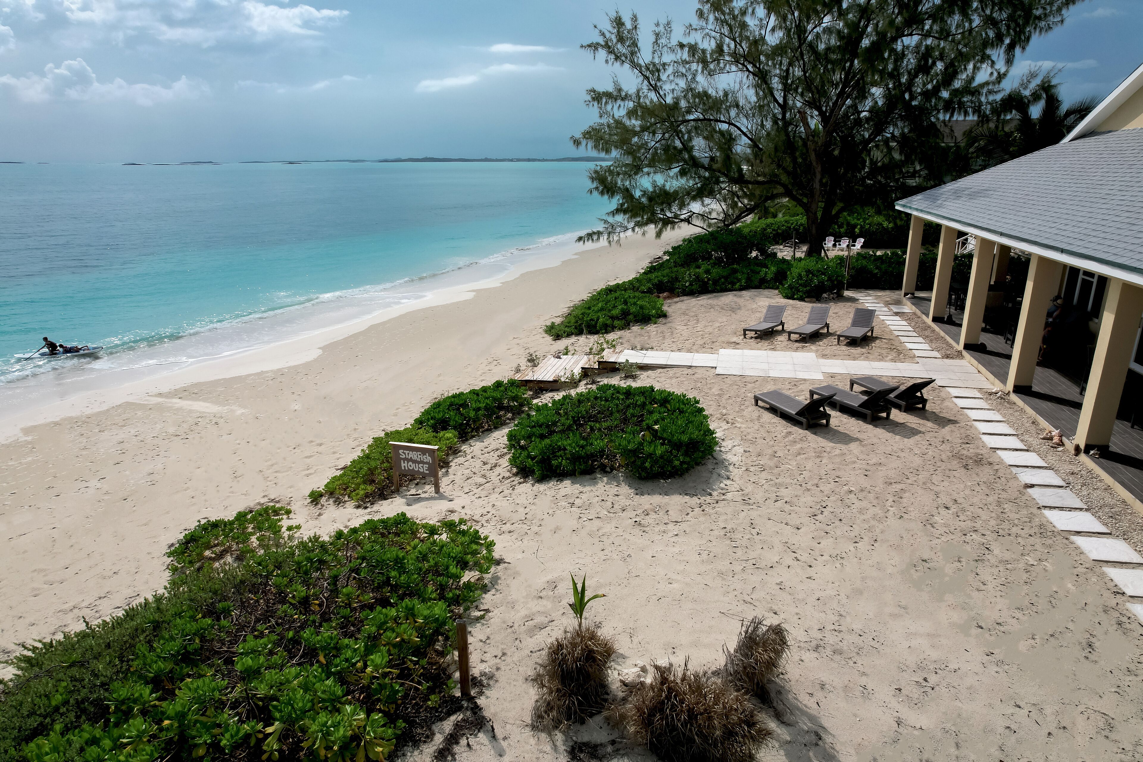 A beachside view with a covered area featuring lounge chairs and a sign labeled "Stargrove House".