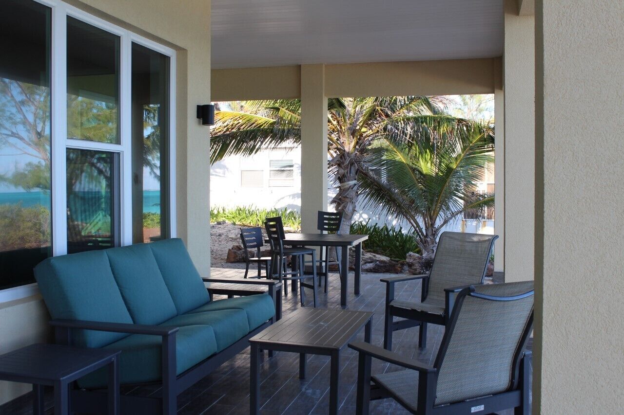 An outdoor patio with a sofa, chairs, and tables, surrounded by palm trees and a view of the ocean.