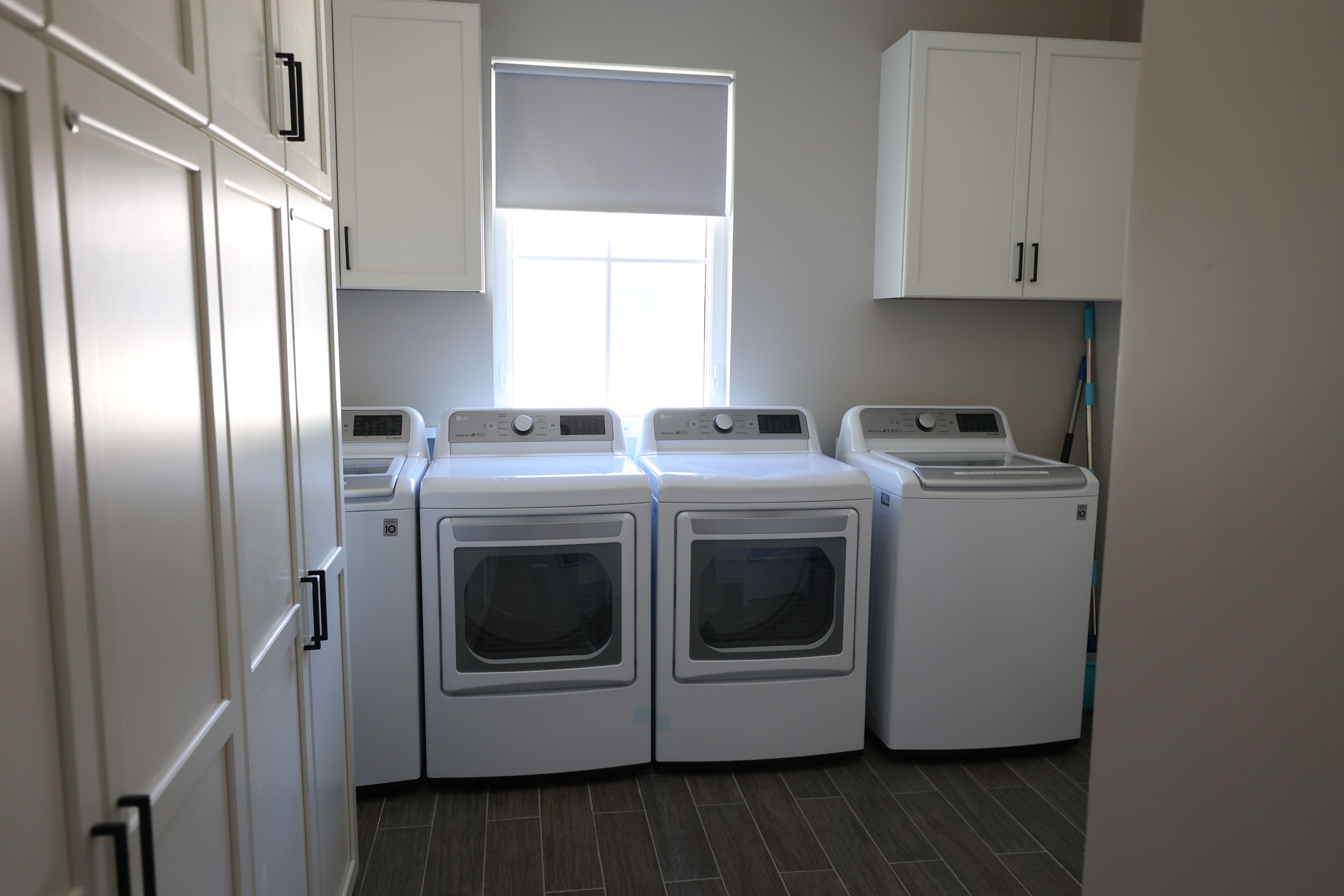 A laundry room with four front-loading washing machines, a window with blinds, and white cabinets.