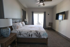 Une chambre à coucher comprenant un lit, une table de chevet, une lampe, un ventilateur de plafond, un téléviseur fixé au mur et une vue sur l’extérieur par une fenêtre.